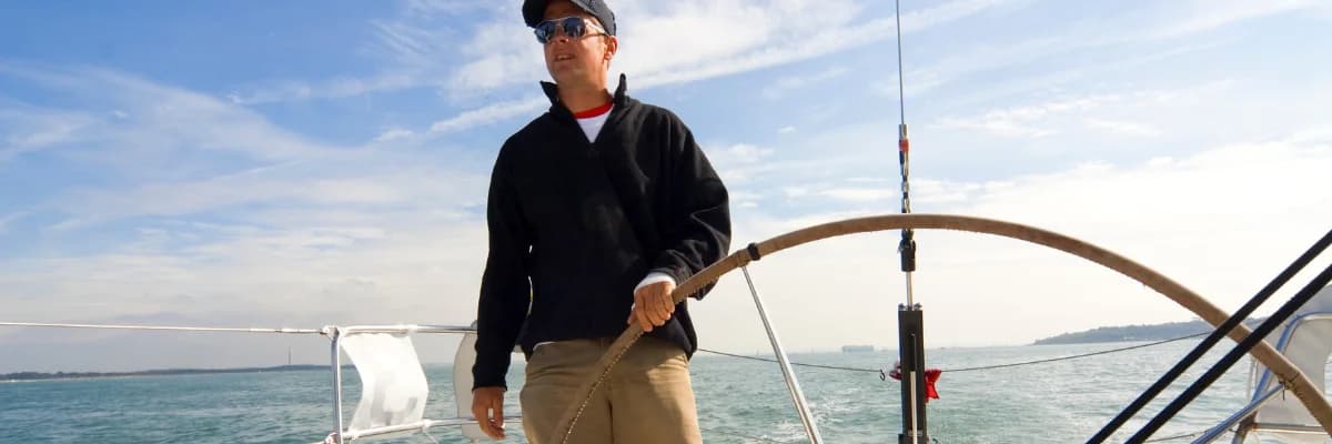 A sailor in a dark jacket steering from the wheel of a sailboat under way